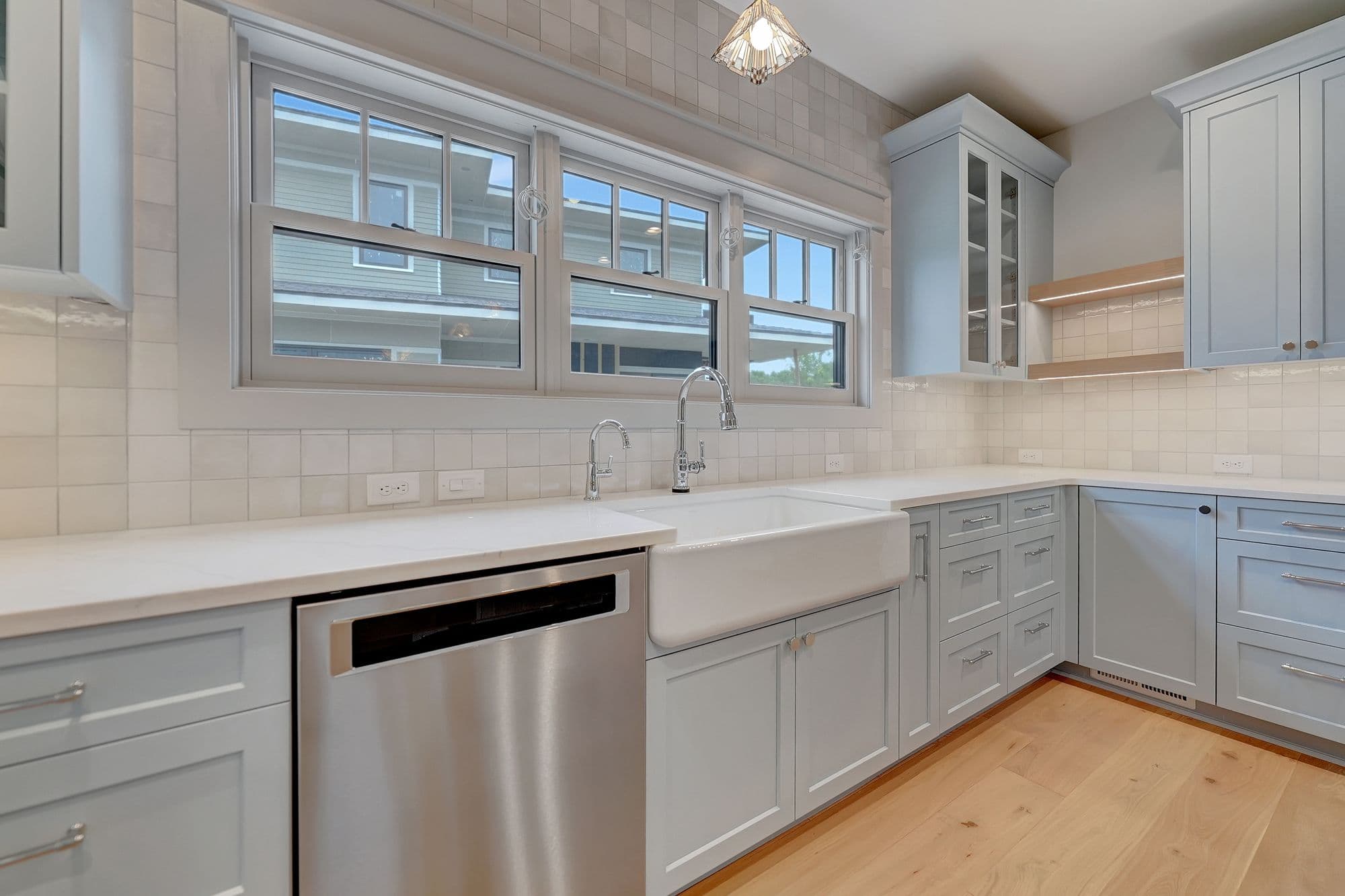 Bright kitchen with light blue shaker cabinets, white countertops and backsplash, and stainless steel appliances. A farmhouse sink is centered under a large window.