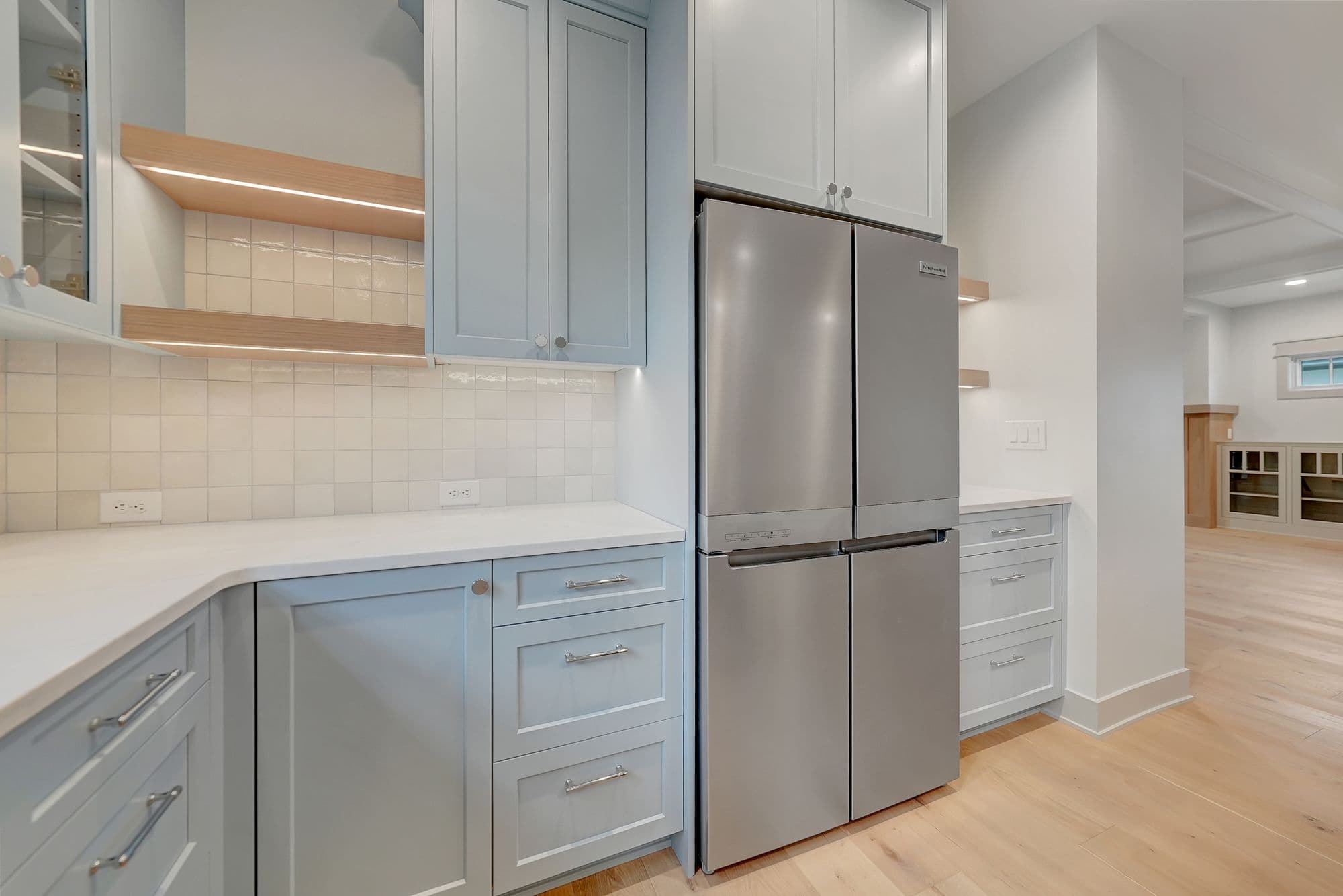 Bright kitchen featuring light blue cabinetry, white countertops, and stainless steel appliances. Open shelving with integrated lighting and a white tile backsplash complement the space.