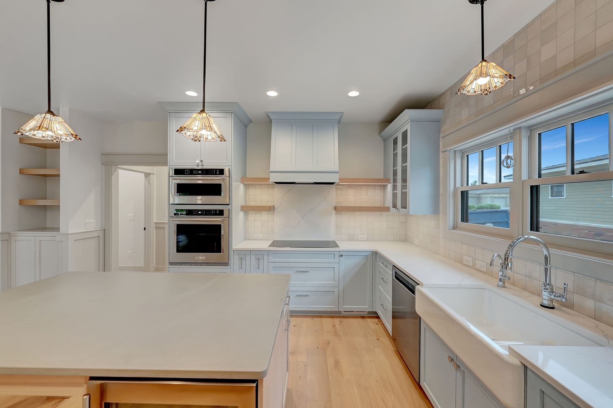 Bright, modern kitchen with light blue cabinets, white countertops, and stainless steel appliances. Features a large center island with light wood accents and three pendant lights above.