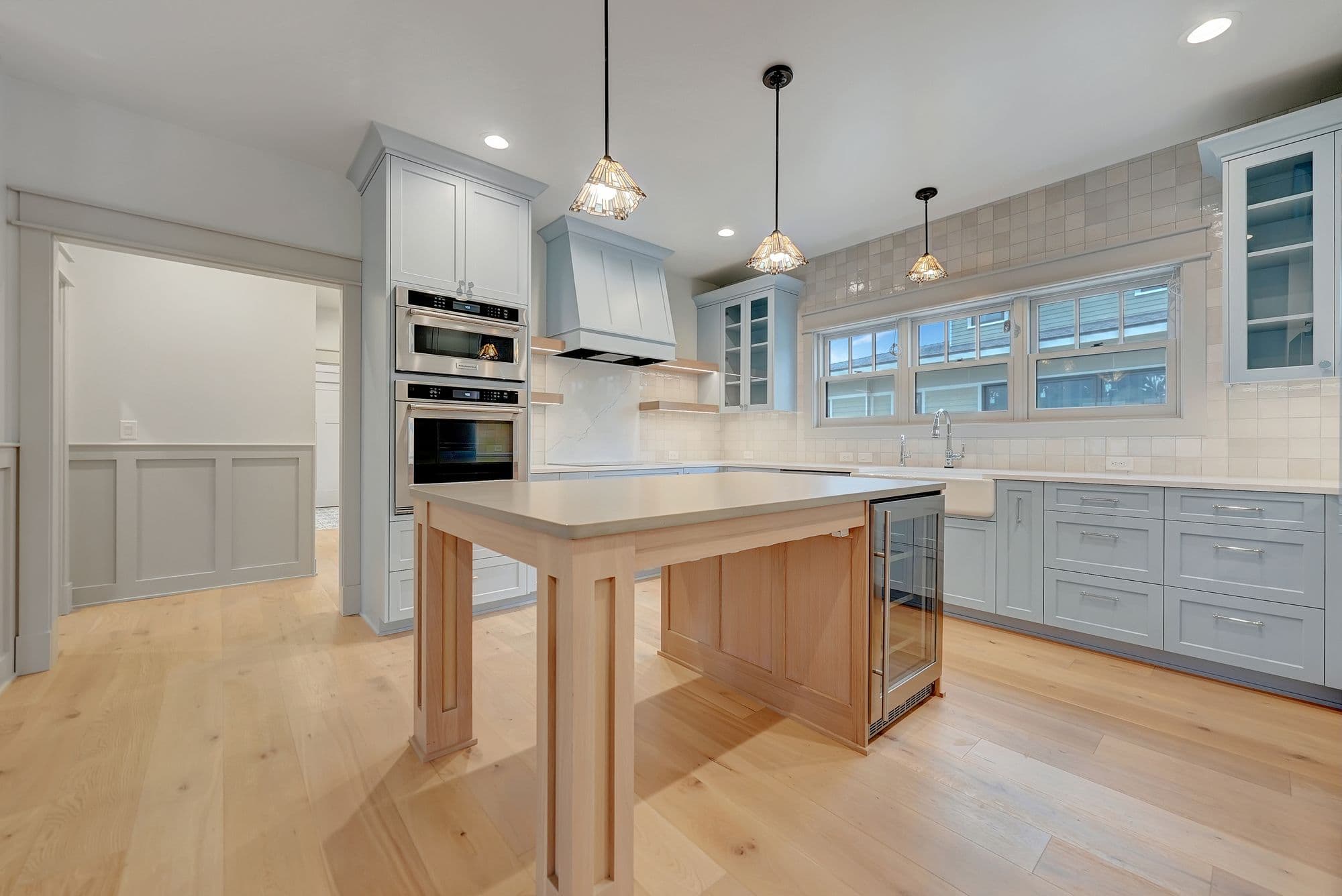 Spacious kitchen featuring blue-gray cabinetry, a light wood island with a beverage fridge, and stainless steel appliances. Natural light pours in through the window above the sink, illuminating the light hardwood flooring.