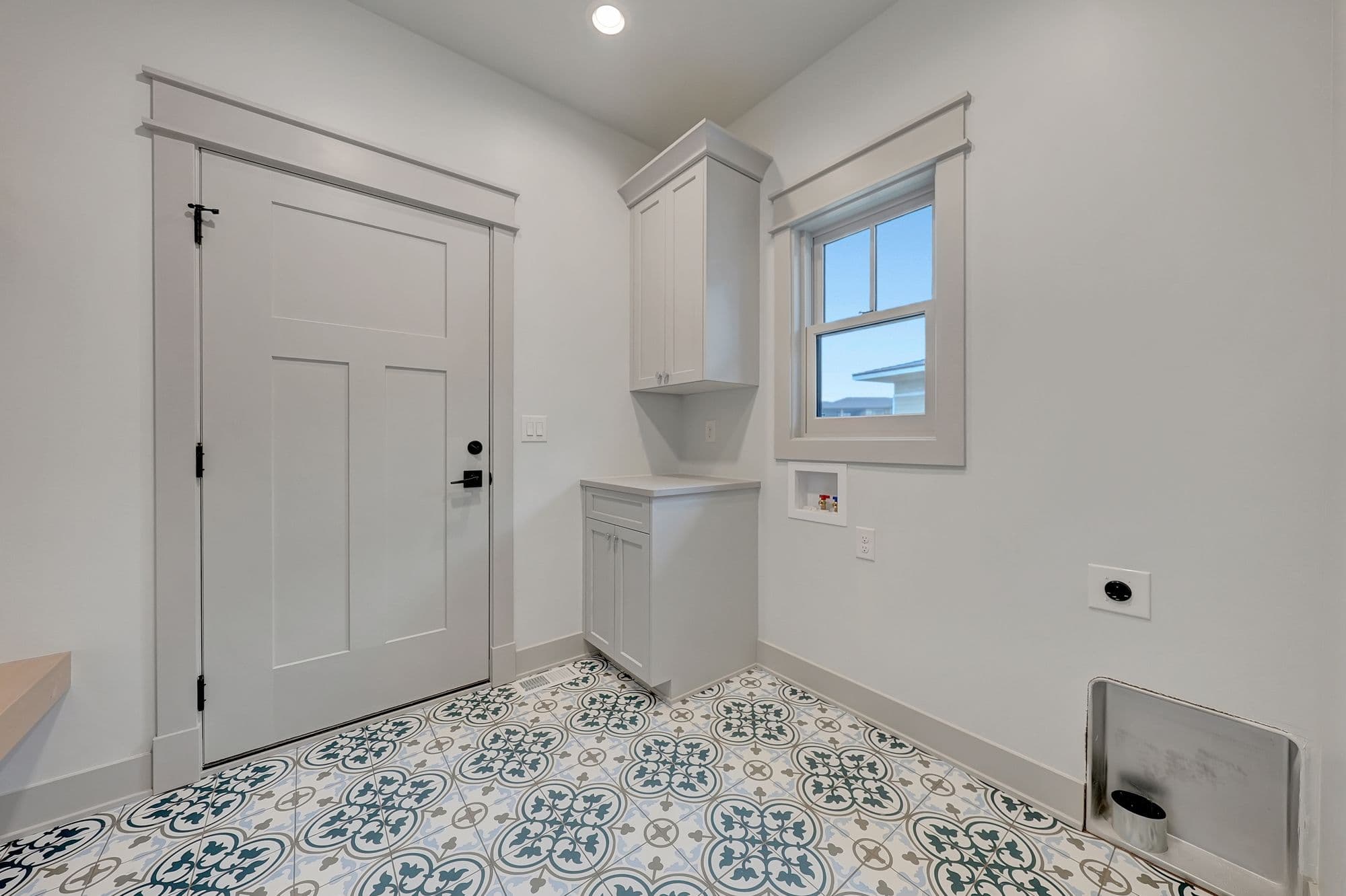 This laundry room features patterned tile flooring with teal and gray accents, light gray cabinetry with storage, and a window. The room has white walls and trim, with a closed door on one side and a laundry hookup on the other.