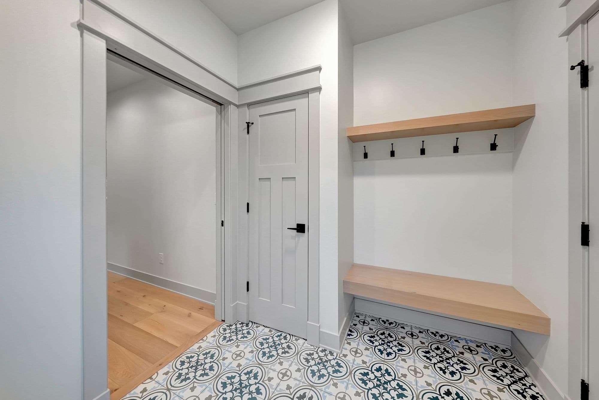 Entryway features decorative patterned floor tiles, white walls, and a built-in bench with coat hooks. A door leads to a closet or storage space with wood flooring.