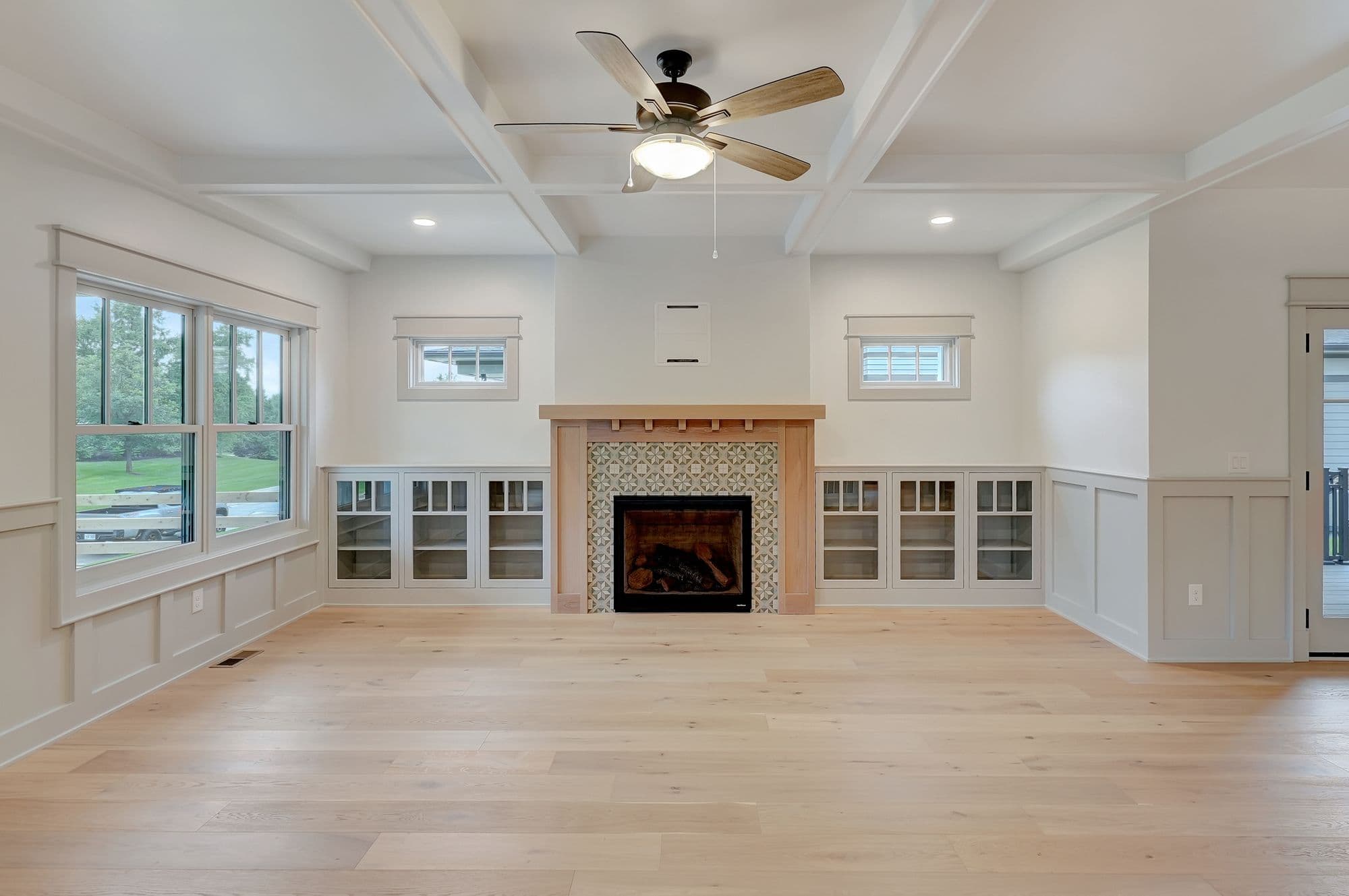 Bright living room featuring light hardwood flooring, white walls with wainscoting, a coffered ceiling, and a fireplace with decorative tile surround. Built-in cabinetry flanks the fireplace, and natural light streams in through large windows.