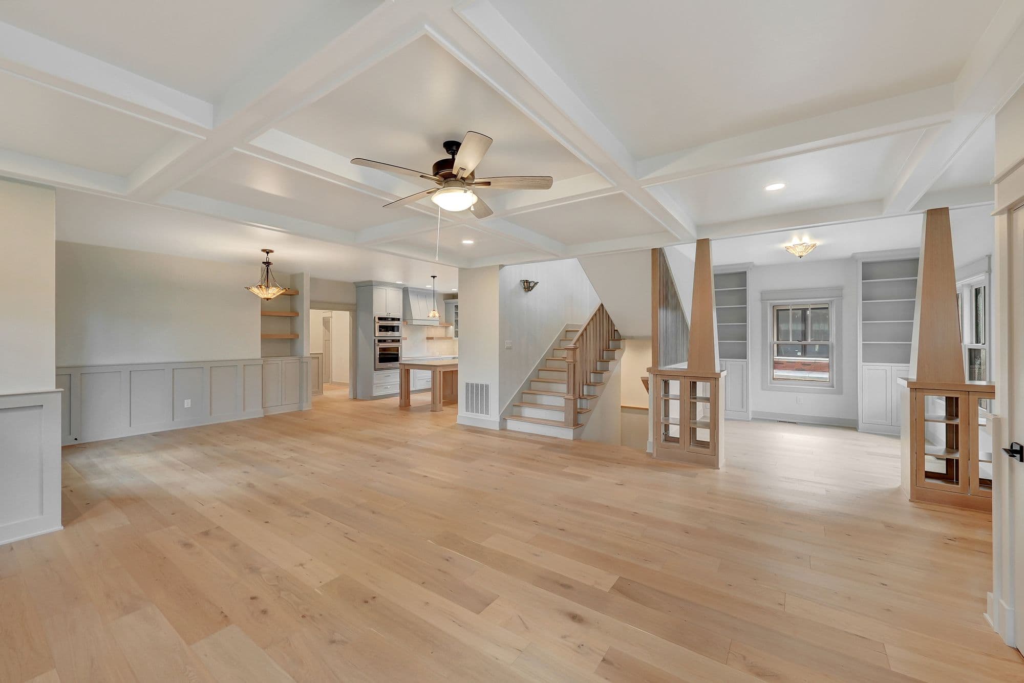 Spacious living room with light hardwood floors and coffered ceilings. Open floor plan leads to a kitchen area and staircase to another level.