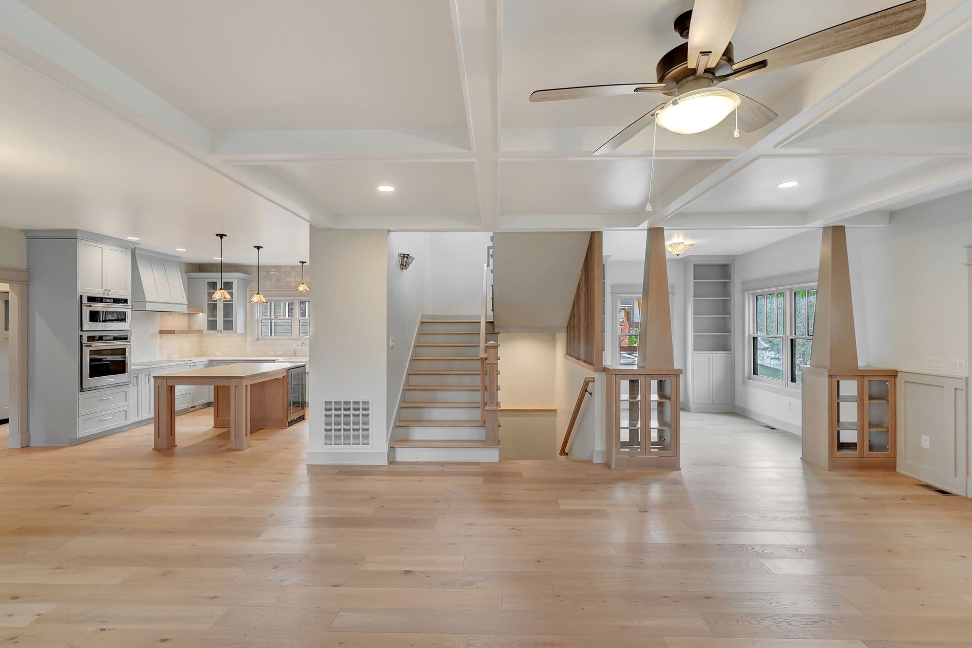 Open-concept living room and kitchen with hardwood floors and coffered ceilings. A wooden staircase leads to another level, and built-in cabinets provide ample storage space.