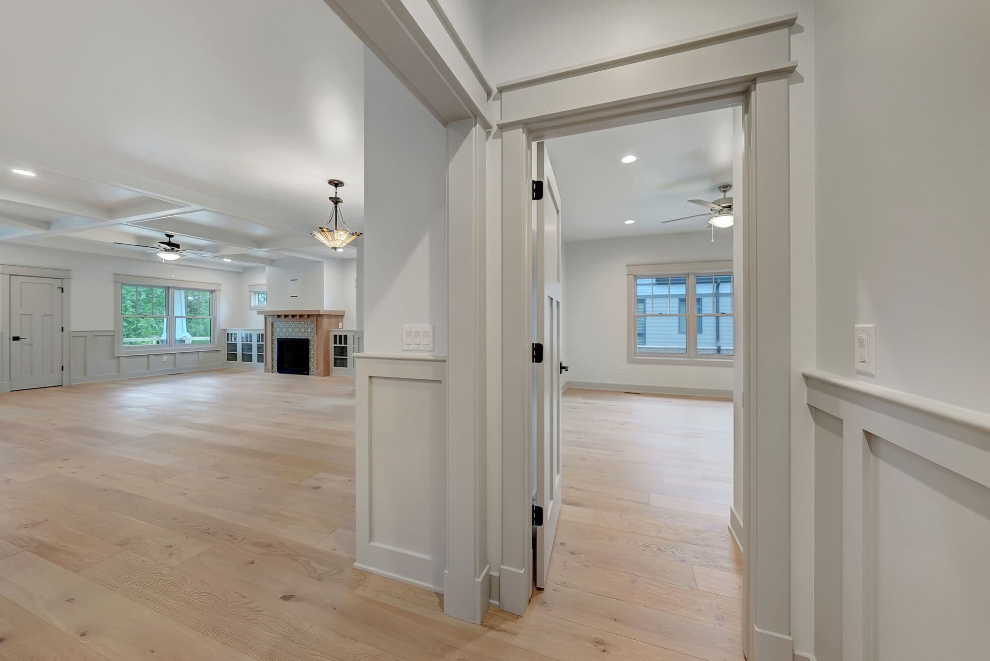 Interior view showcases a light-filled open floor plan with light hardwood floors, transitioning from a living room with a fireplace and built-in shelves to a bedroom through a partially open door. The rooms feature white trim and neutral wall colors.