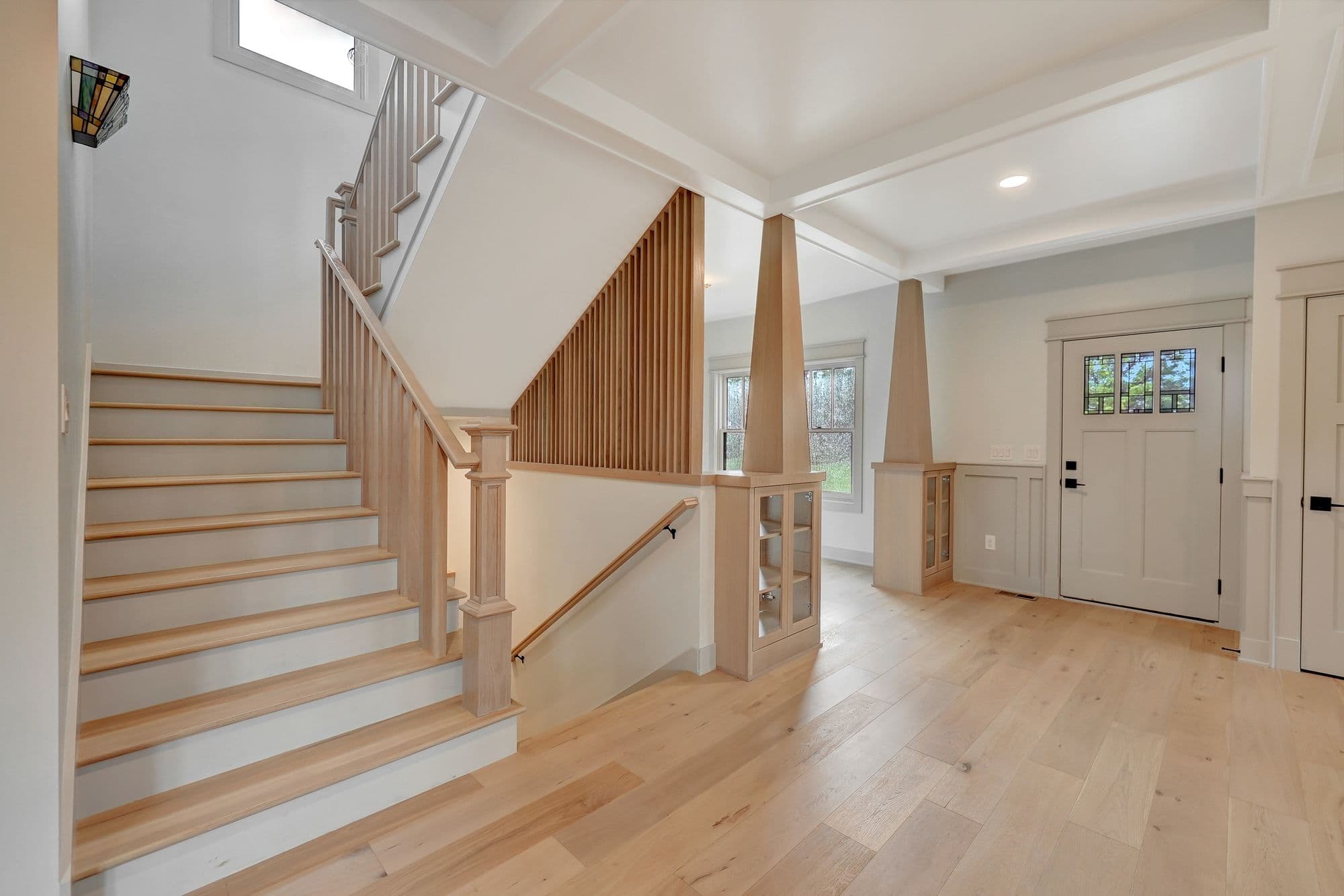 Bright entryway with light wood flooring, a staircase with wood treads and white risers, and a front door with decorative glass panels. The space features built-in cabinets, wood trim, and white walls.