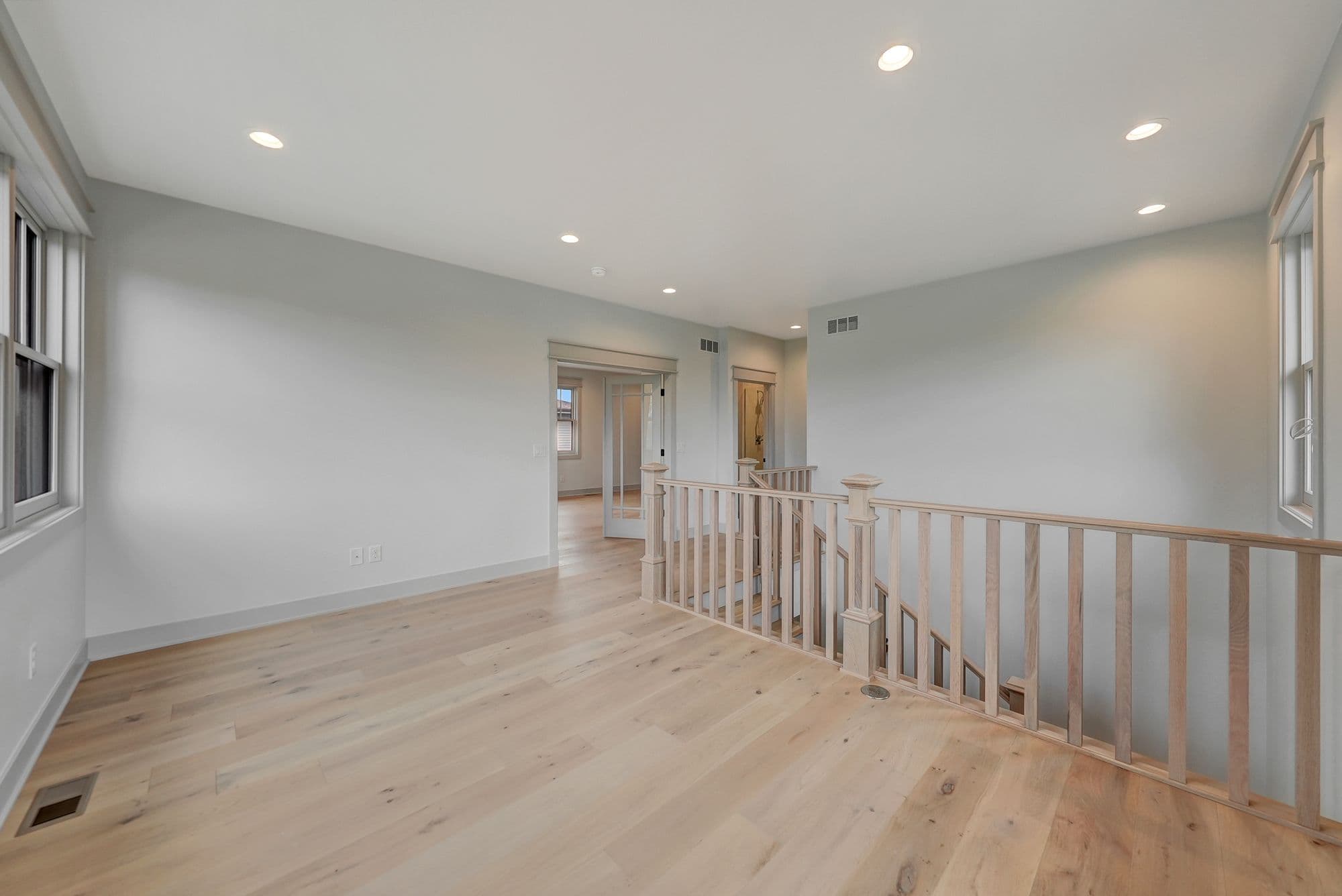 Bright, open landing area features hardwood floors and a wooden railing overlooking the stairs. Two doorways lead to other rooms, and natural light filters through a window.
