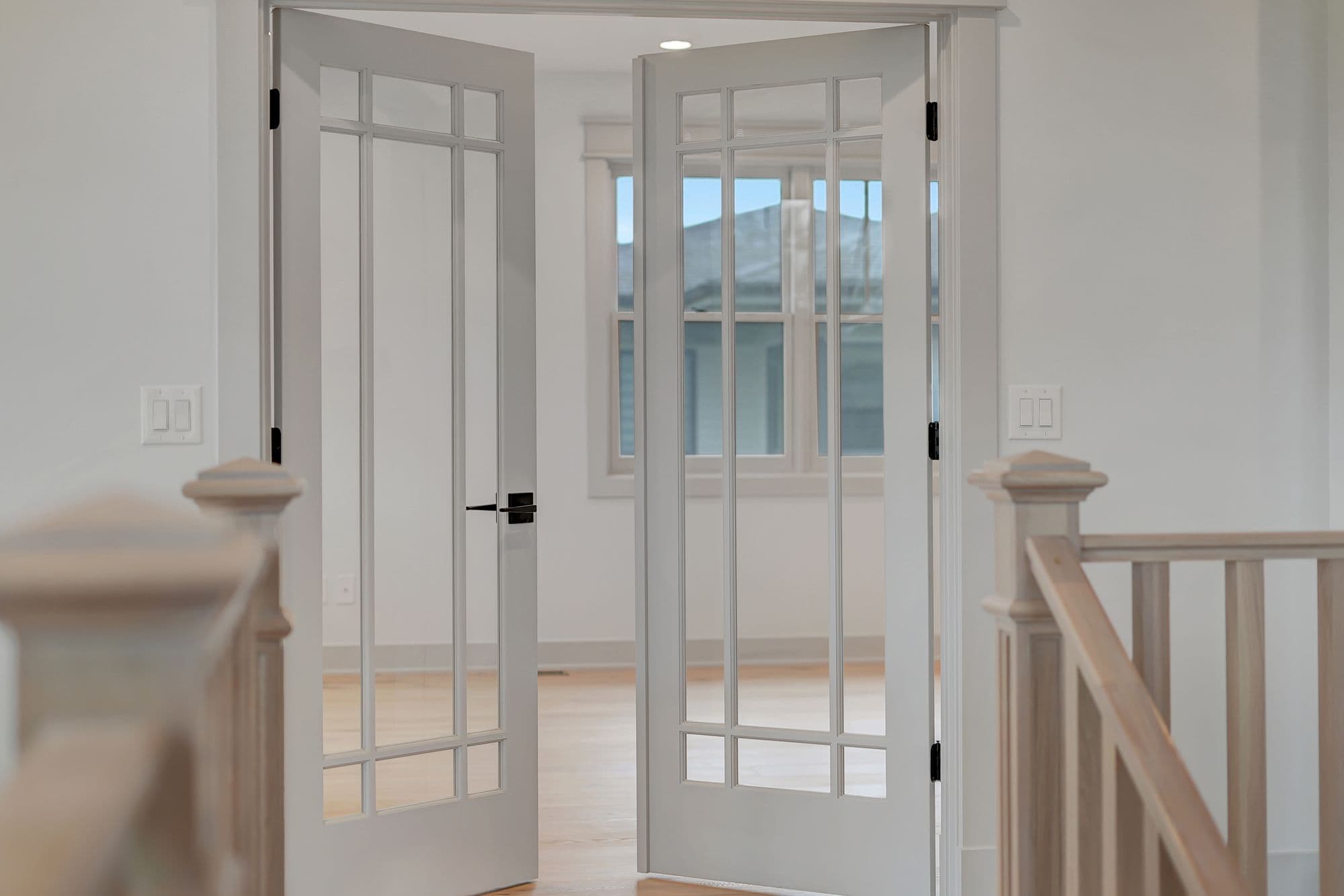 Interior view through open French doors with glass panes. The view shows a room with light wood floors and a window.