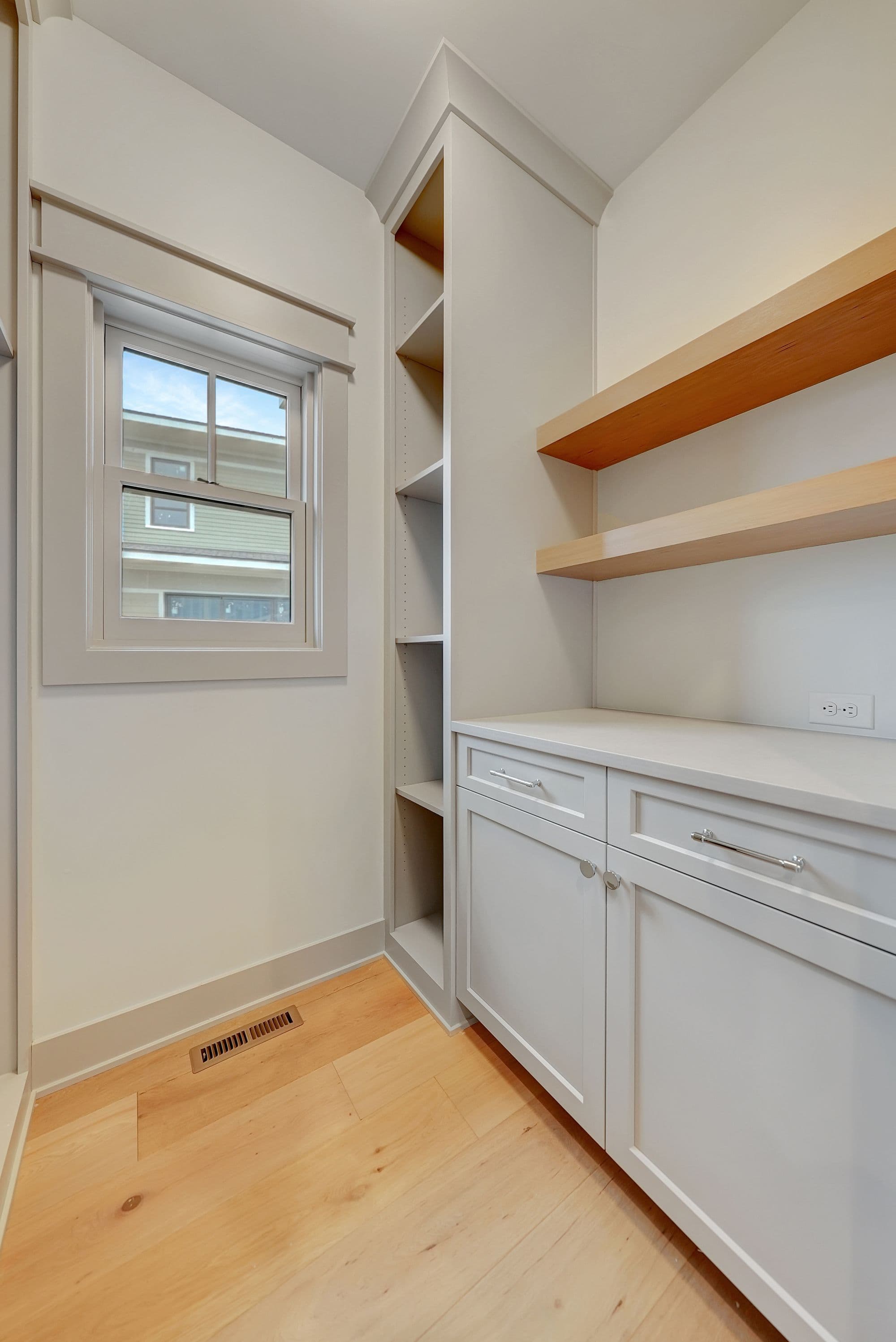 Bright pantry area with light wood flooring, gray built-in cabinetry, and open wood shelves. A window allows natural light into the space.