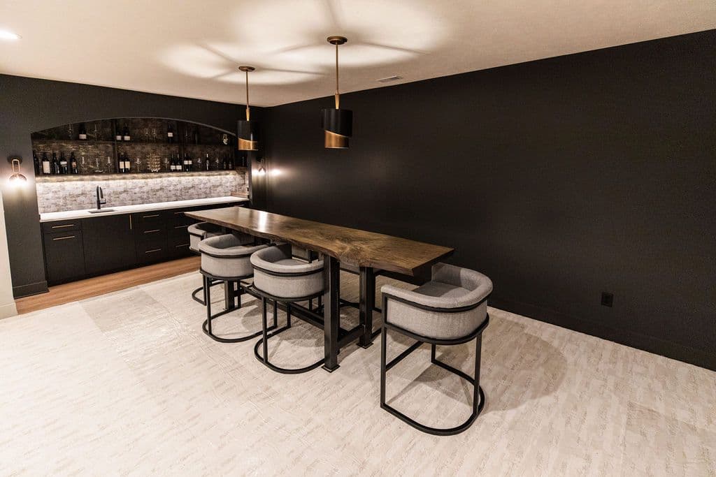 Modern basement bar area features a long live edge wood countertop with black metal base and gray upholstered barstools. The bar includes a sink, cabinets, and wine rack built into an arched alcove with dark gray walls and light tile floor.