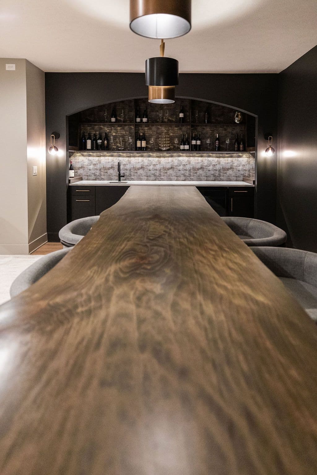 Contemporary bar area features a live edge bar top, dark cabinetry, and a backsplash with under-cabinet lighting. The back bar includes open shelving with bottles and glassware, lit by sconces flanking the area.