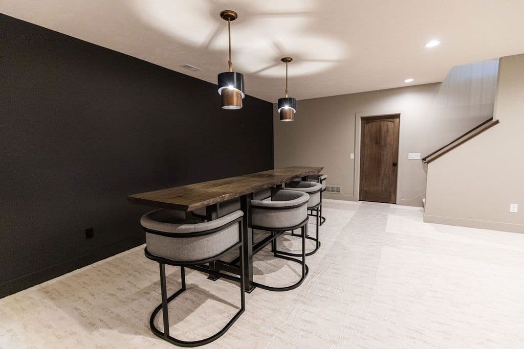 Finished basement area with a long wooden bar and four gray upholstered bar stools. The room has a dark accent wall, light-colored patterned flooring, and modern pendant lighting.