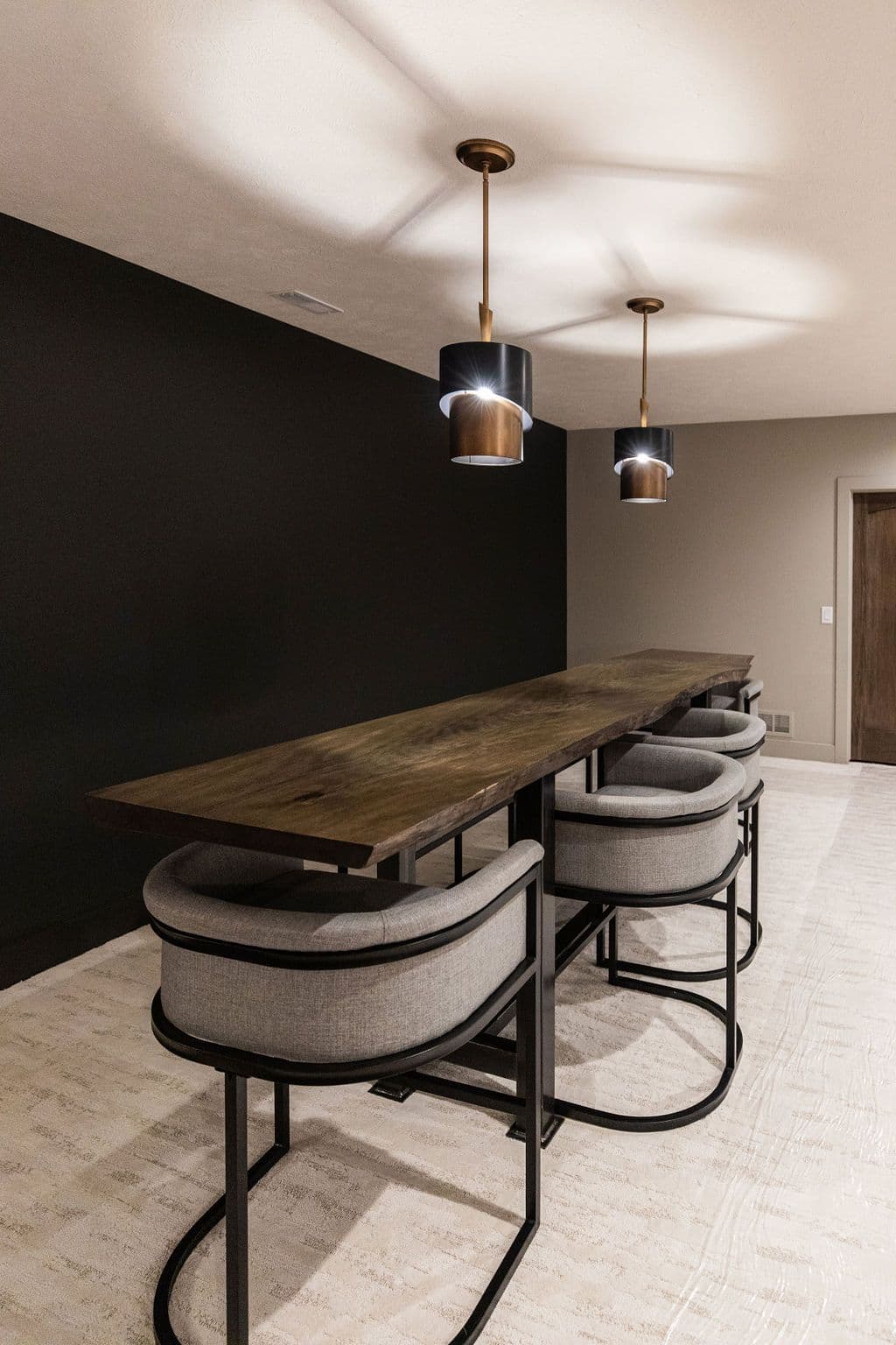 Modern basement bar area with a live edge wooden countertop, steel-framed bar stools upholstered in gray fabric, and pendant lighting above. The space is defined by a contrasting black wall and light flooring.