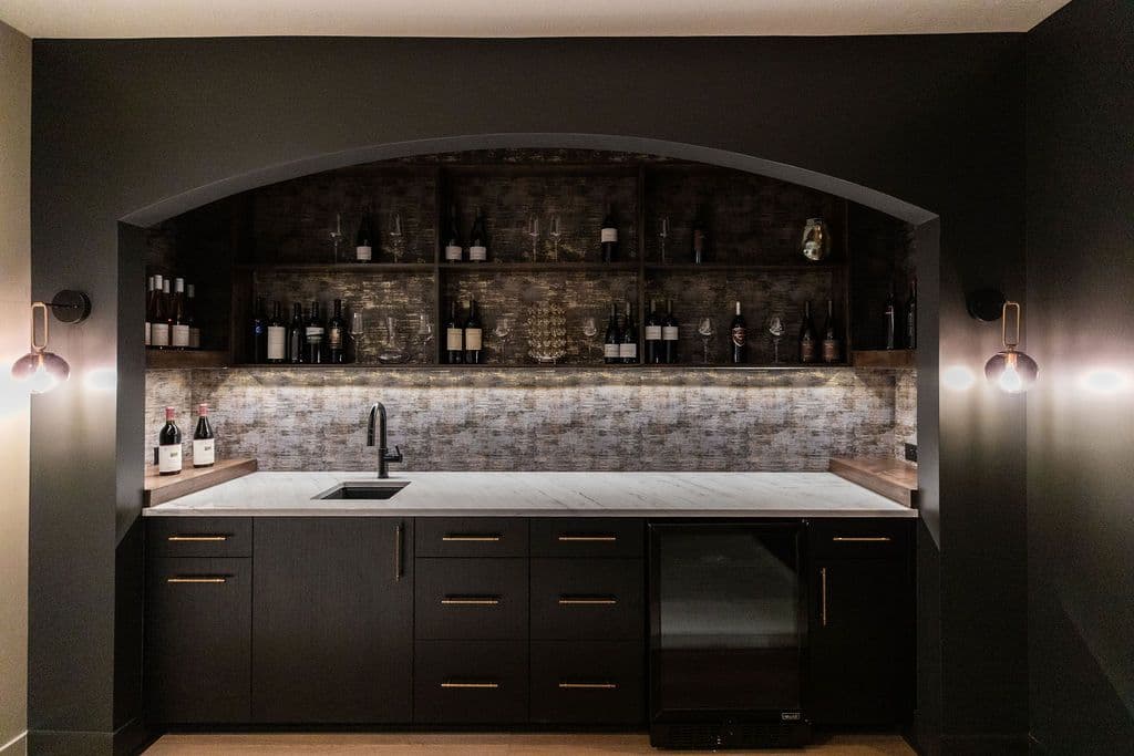 Dark home bar area featuring built-in shelving with wine storage, marble countertops, and dark cabinets. Wall sconces provide lighting on either side of the bar.