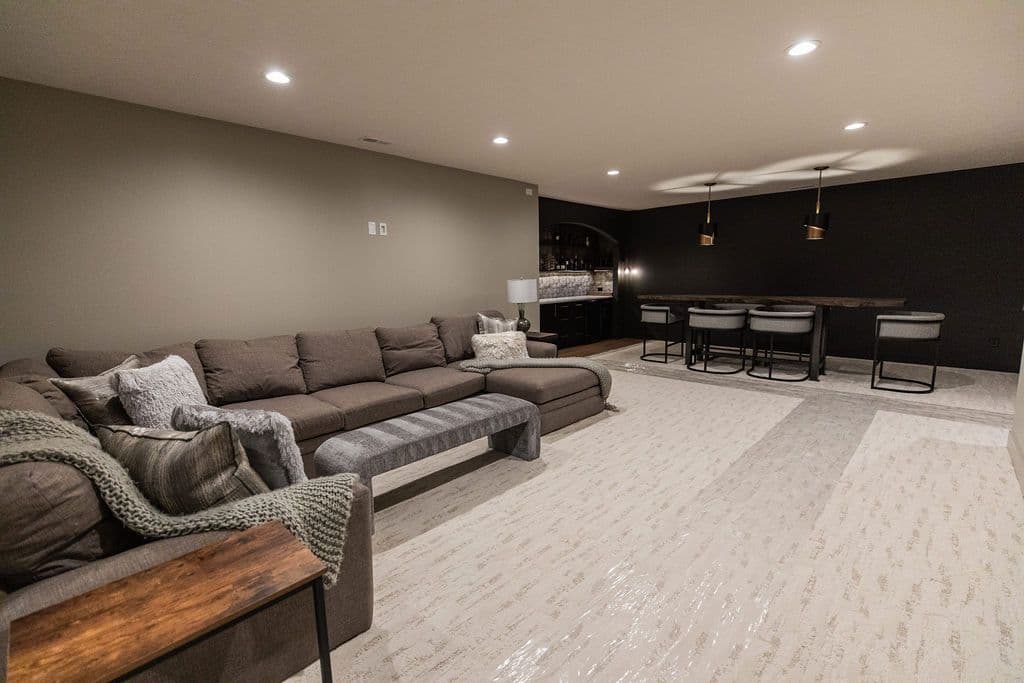 Finished basement space features a large sectional couch, long bench, and a bar area with seating. The space is lit by recessed lighting and the walls are painted in contrasting dark and light tones.