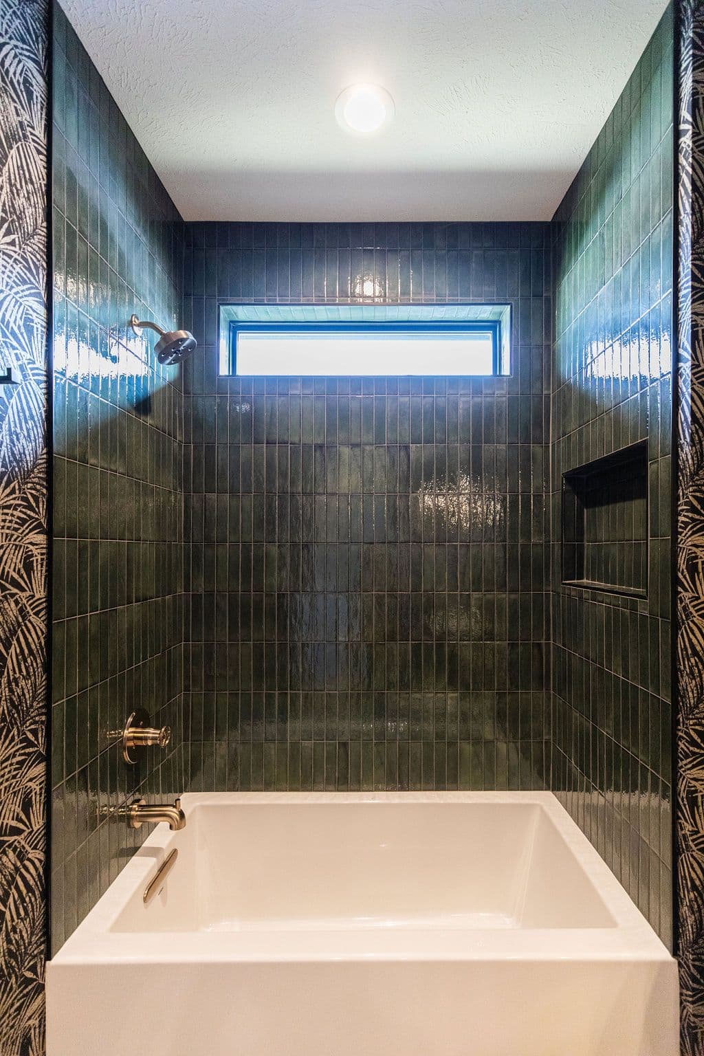 Bathroom features a modern tub with a rectangular window and olive green vertical tiling surrounding the tub and continuing to the walls. One wall is accented with black and cream leaf-patterned wallpaper.