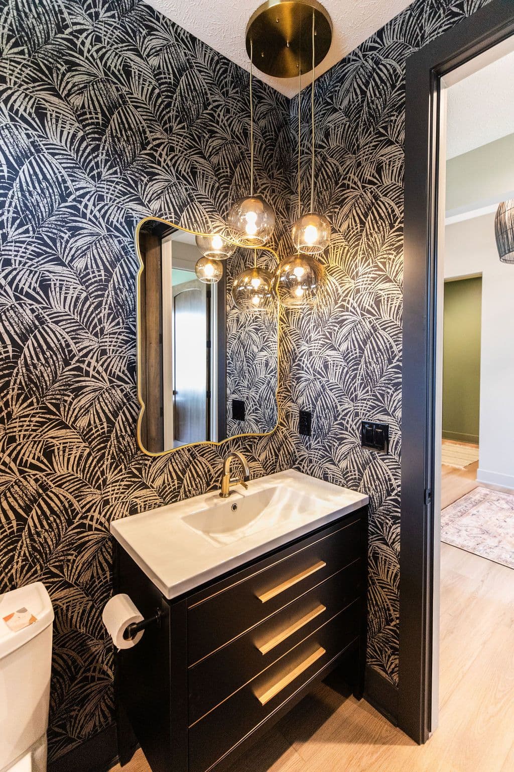 Powder room featuring black and gold palm leaf wallpaper, gold-framed mirror, and a black vanity with gold hardware. Multi-bulb gold light fixture hanging from the ceiling, with a toilet visible to the left.