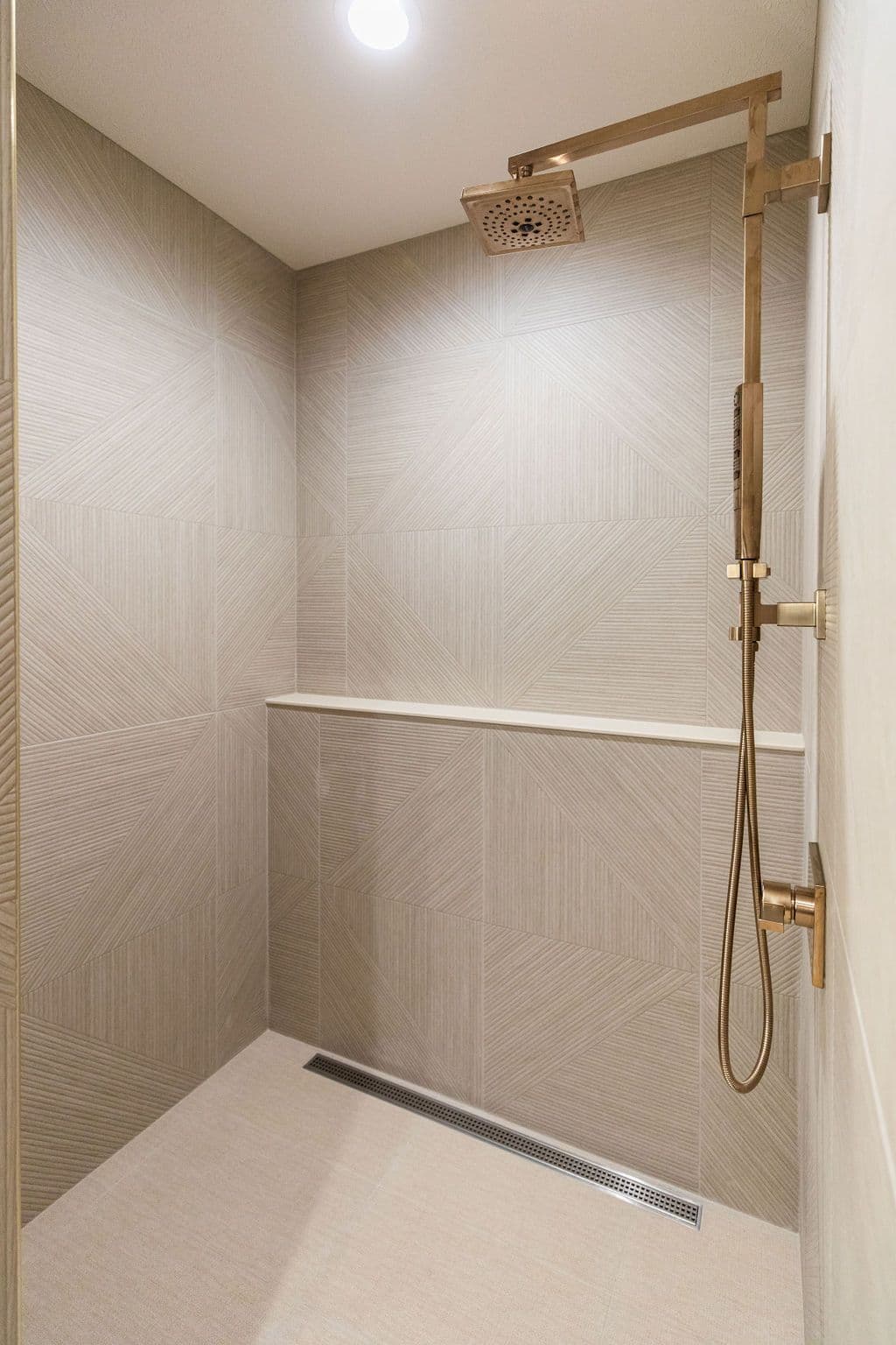 Modern shower with geometric patterned tiles and a bronze shower head and fixtures. The shower has a built-in shelf and a linear drain.