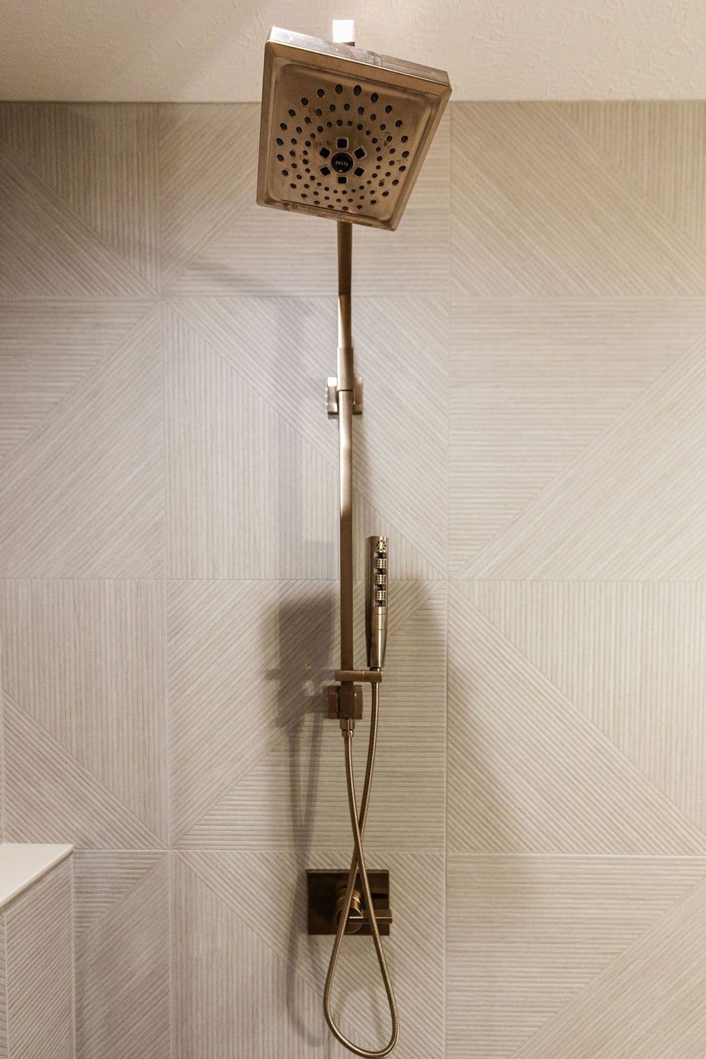 Bathroom with Delta shower head and handheld wand, on tile wall with geometric pattern. The colors are light and neutral with brass fixtures.