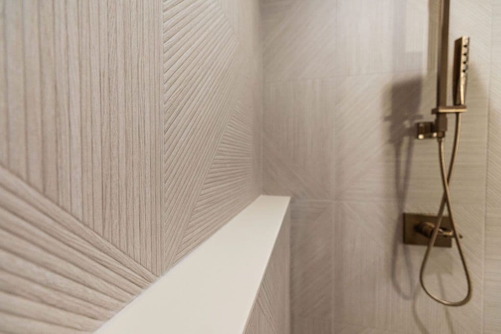 A modern bathroom features geometrically patterned beige wall tiles with a narrow white shelf and a brushed gold shower head. The wall tiling has a textured appearance that mimics a wood grain.