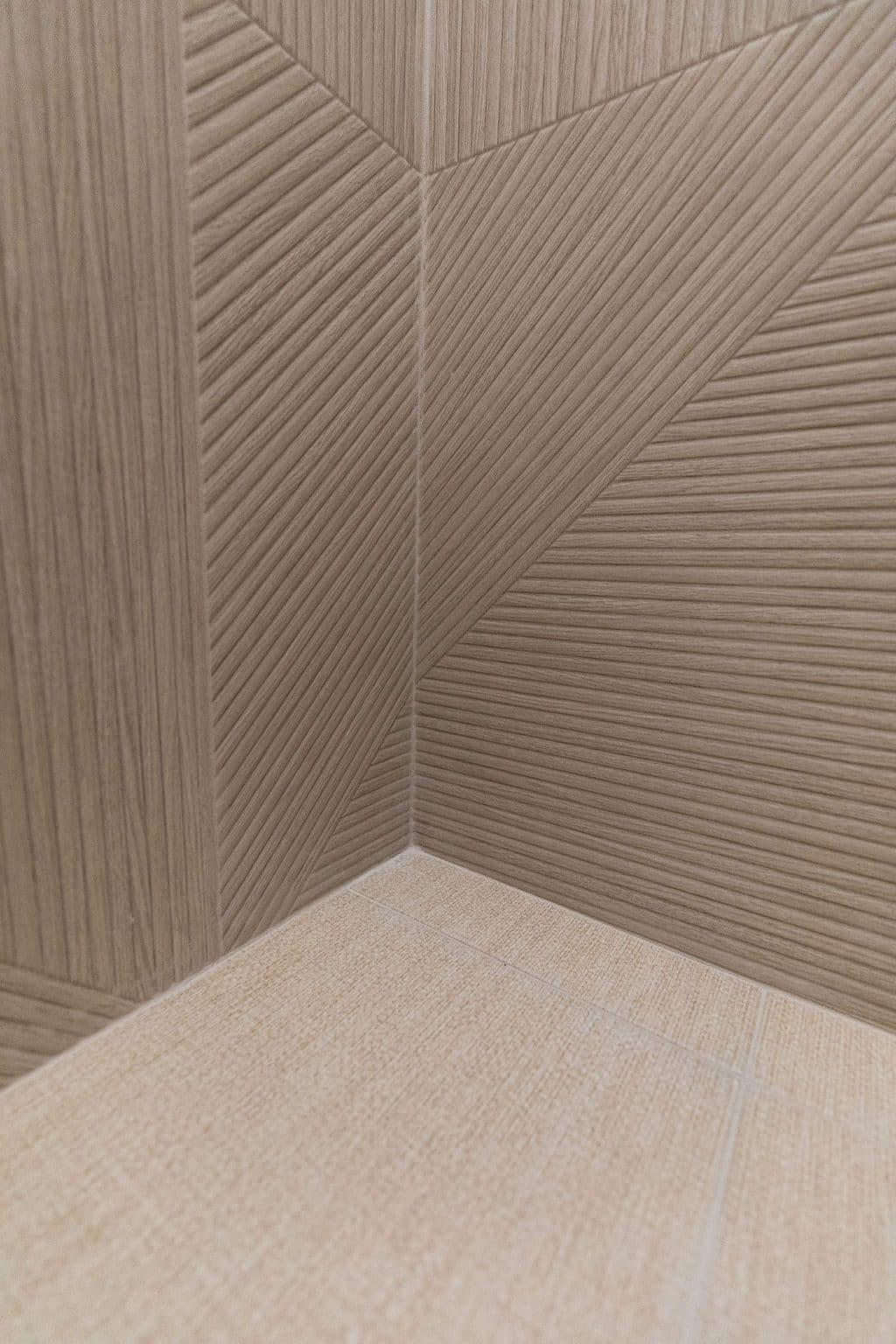 Corner of a room featuring textured tile walls with a geometric pattern and matching textured floor tiles in a neutral color palette. The wall tiles are vertically oriented on the left side and angled on the right, creating a visually dynamic space.