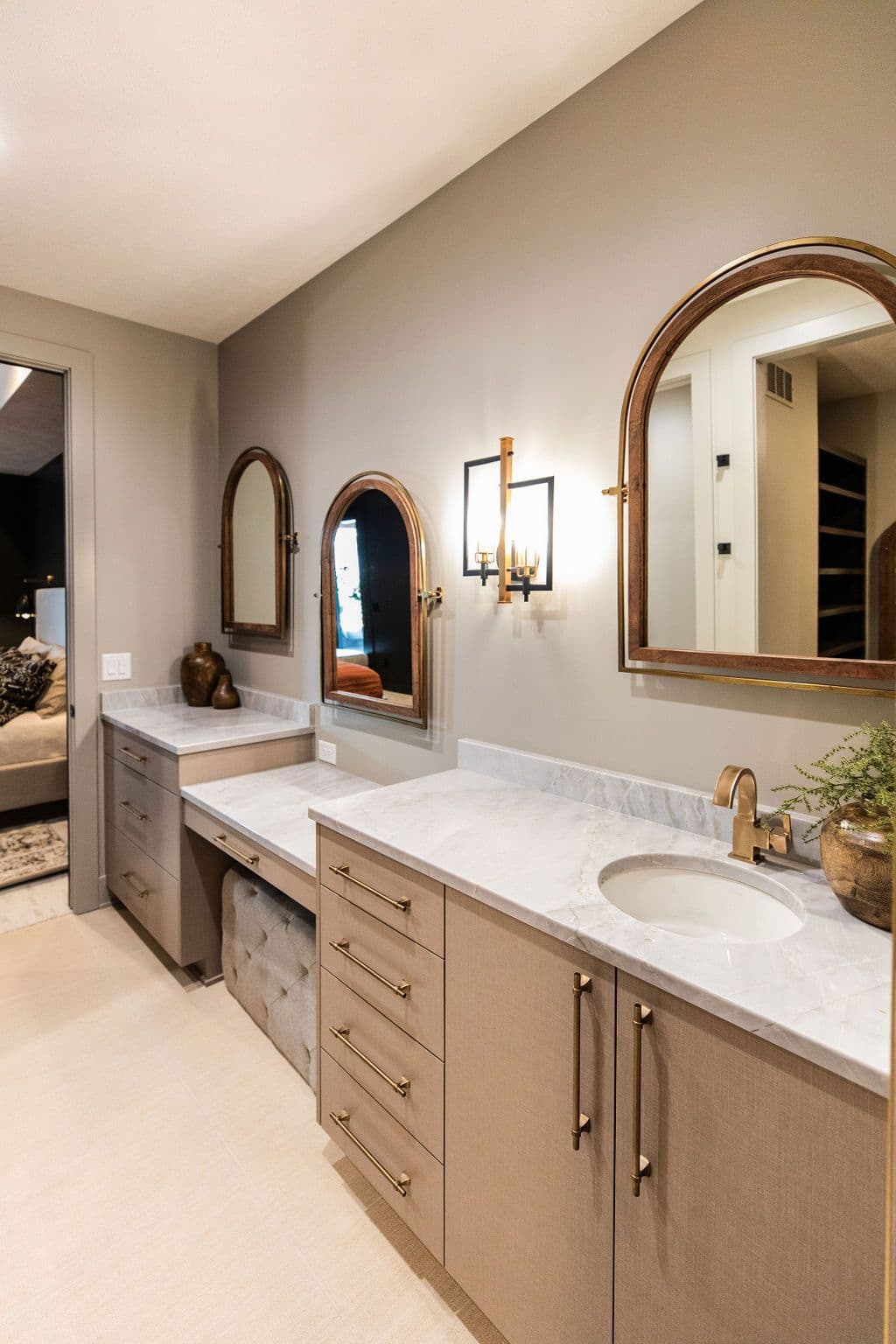 Bathroom vanity area with light brown cabinets, marble countertops, and arched mirrors. Features brass fixtures and a seating area between the two vanity spaces.
