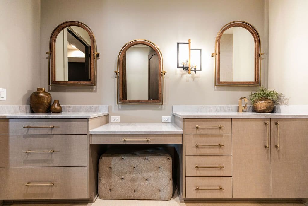 Bathroom vanity features three arched wood-framed mirrors over a countertop with neutral-toned cabinets and drawers, accented by gold hardware. A tufted ottoman is positioned beneath the vanity, providing a seating area.