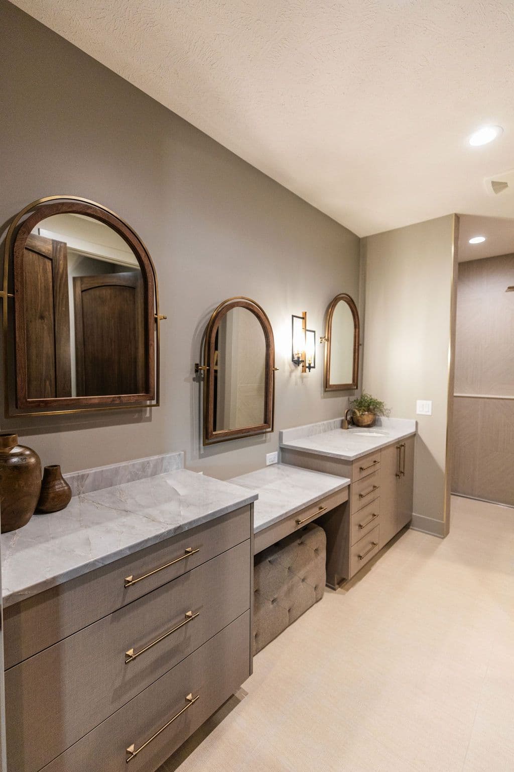 Bathroom vanity area features three arched mirrors, taupe cabinets with gold hardware, and marble countertops. The space also includes a tufted ottoman and tile flooring.