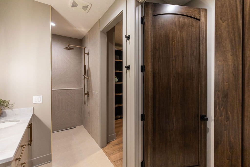Bathroom showcases a modern shower with geometric tile and gold fixtures, a vanity with marble countertop and gold hardware, and a dark wood door leading to a closet or storage area, creating a sleek and functional design.
