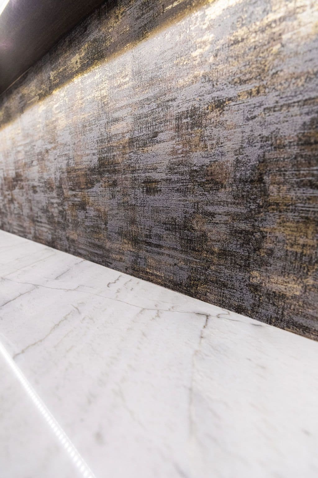 Close up of a marble countertop with light and gold accented wall texture. The countertop is white with gray veining.