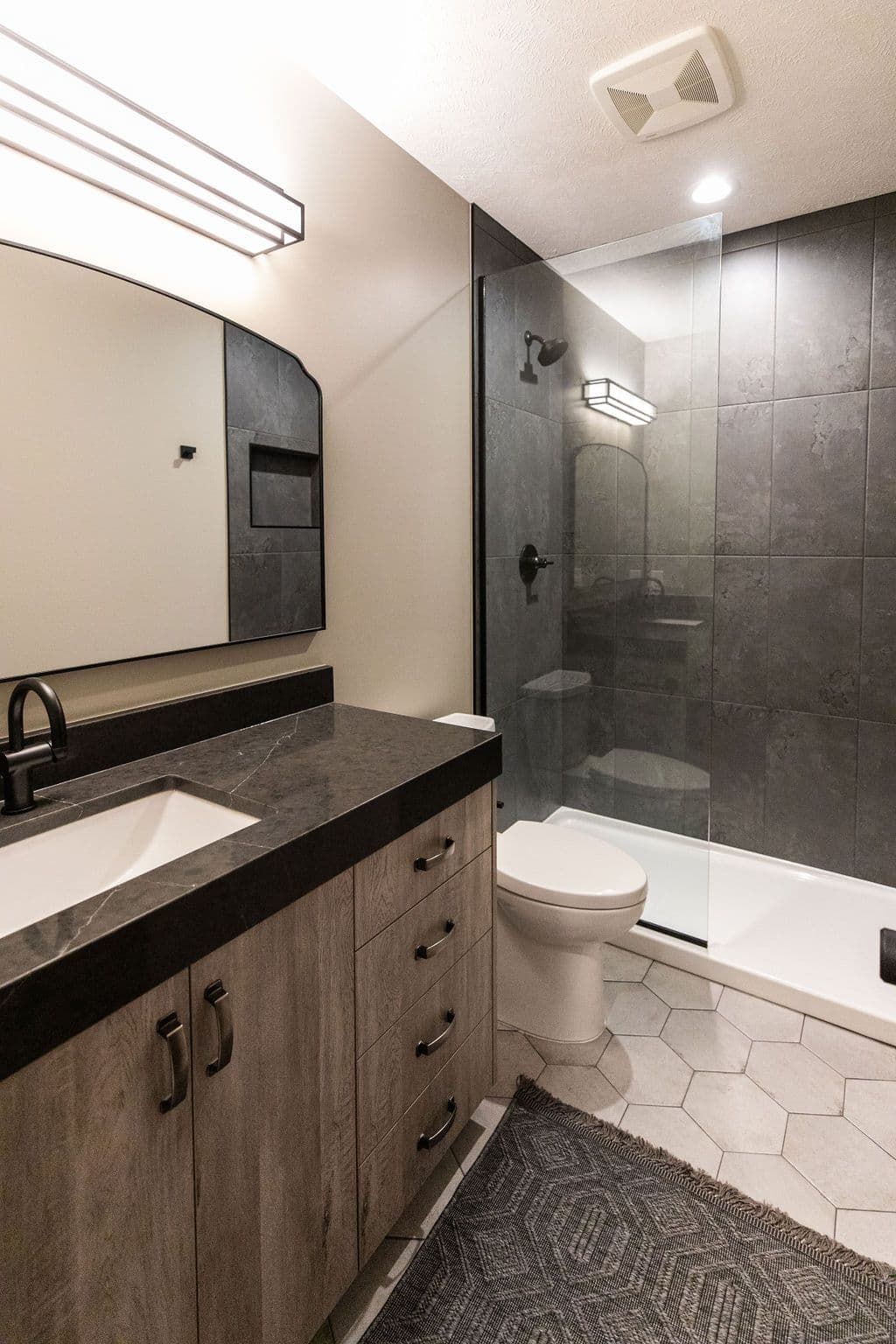 Modern bathroom features a wood-toned vanity with dark countertop, matte black faucet, and a wall-mounted mirror. Walk-in shower is enclosed with glass and has gray tiling, along with gray hexagonal floor tiles and a patterned gray rug.