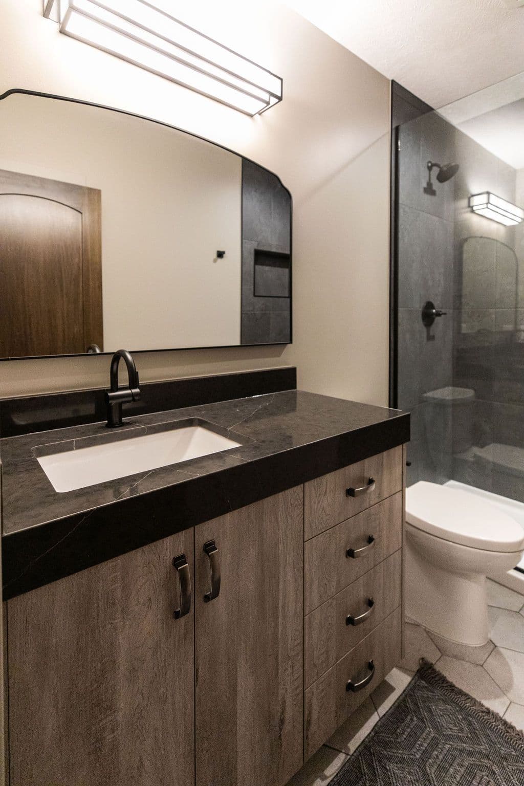 Bathroom interior featuring a vanity with a gray countertop and light wood cabinetry. The space has a modern aesthetic with a black framed mirror and a glass-enclosed shower area.