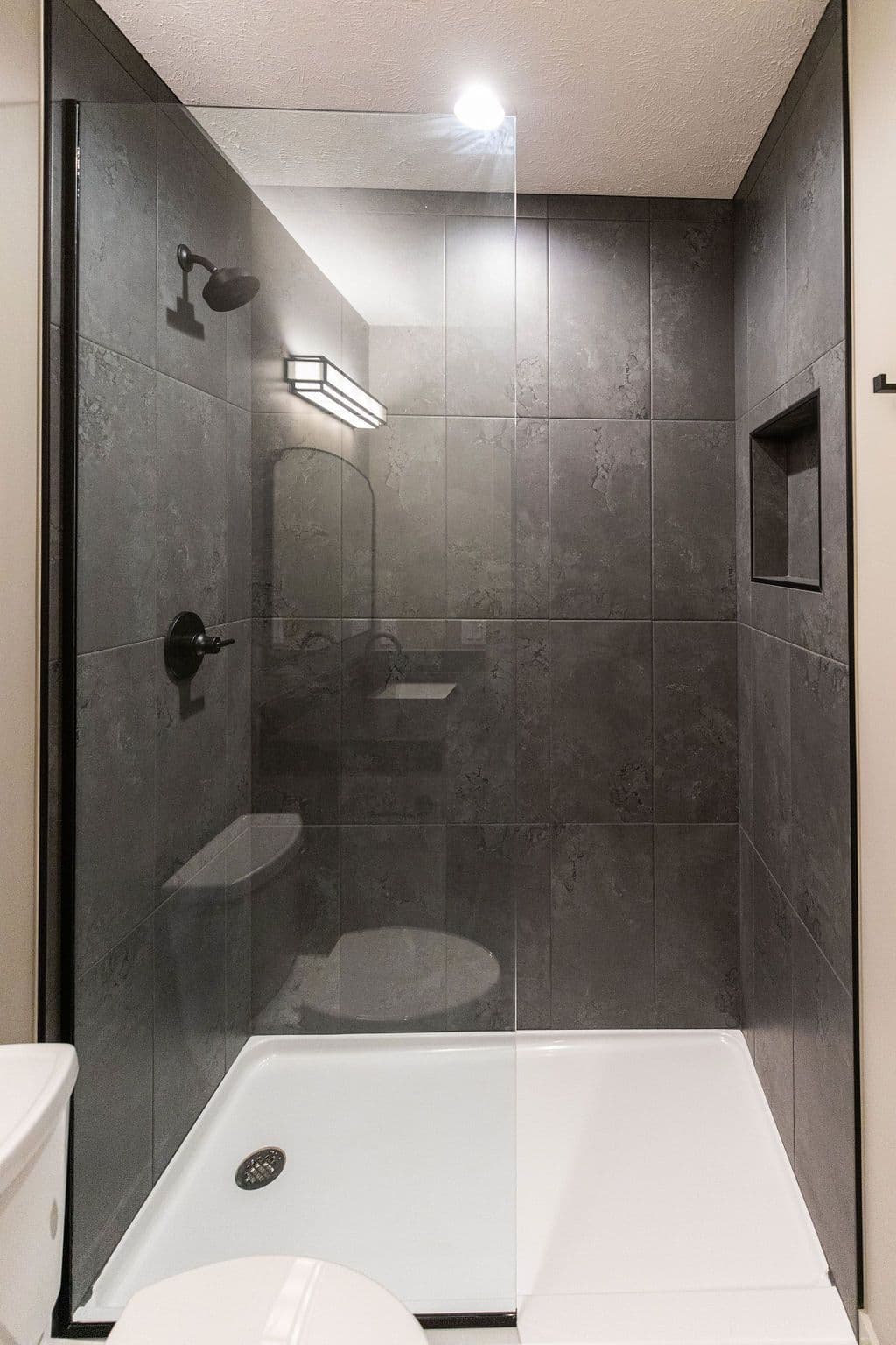 Modern bathroom with large gray tiled walls, black fixtures, and a glass shower enclosure. The shower features a built-in niche and a white shower base.