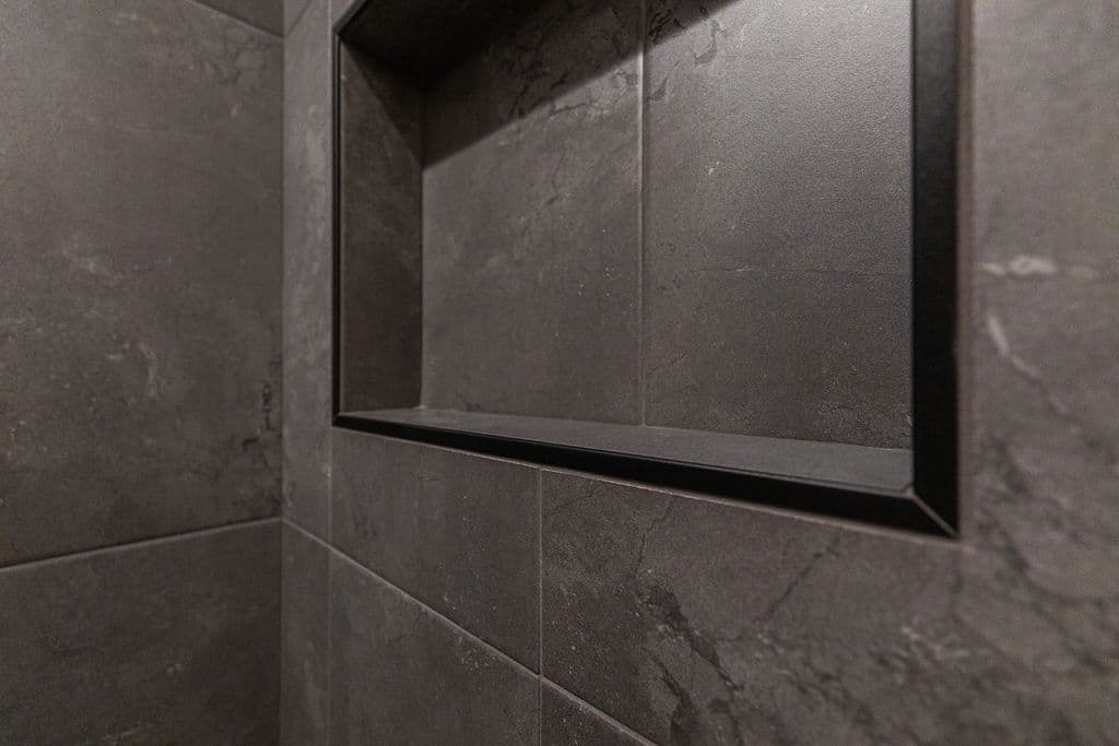 Modern bathroom features large format gray tile walls with a built-in black-framed shower niche. The tile has a textured stone-like appearance.