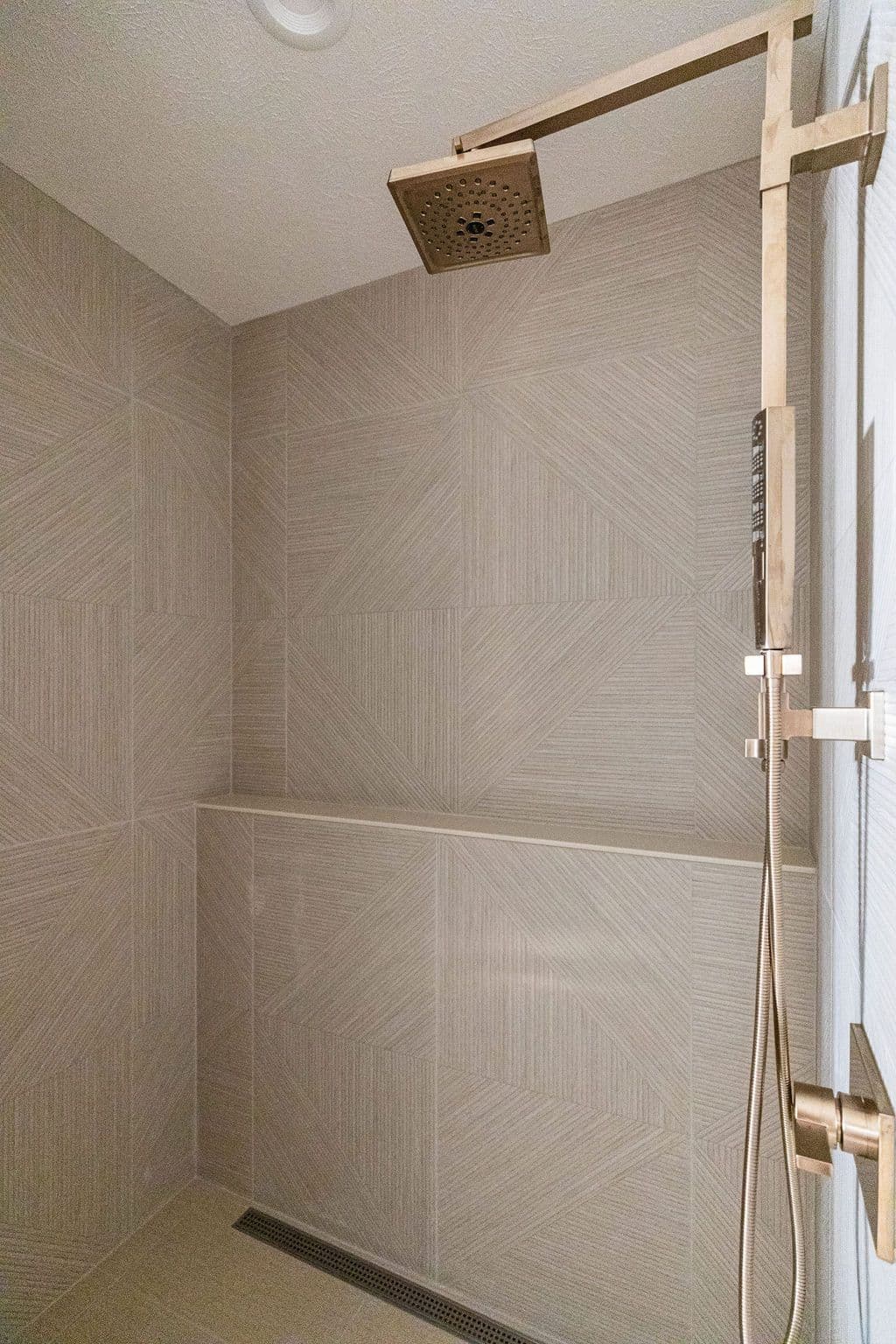 A modern shower area features geometric tile walls in shades of beige, complemented by bronze hardware including a rain shower head and handheld shower fixture. A built-in shelf and linear drain contribute to the clean design.