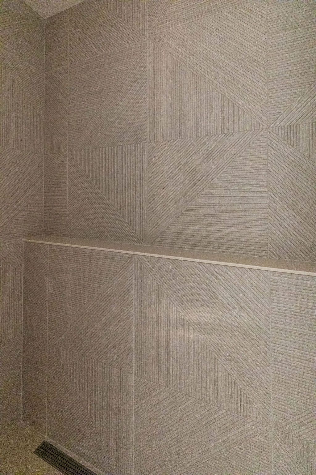 Bathroom showcasing textured geometric tile work with horizontal lines and a built-in shelf. The tiling covers the walls and extends to the floor, with a visible drain.