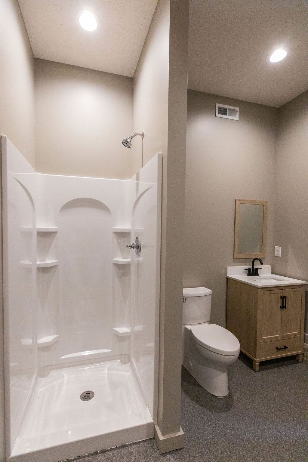 Bathroom with a white shower stall, toilet, and vanity. The walls are a neutral beige color and the floor is a gray speckled material.