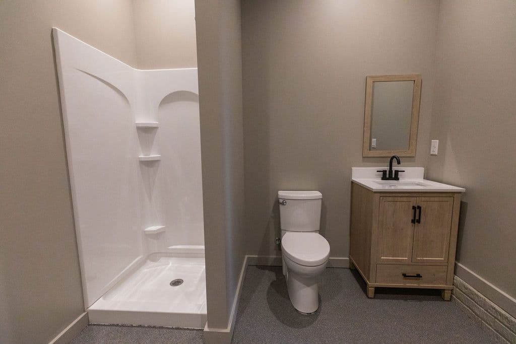 Bathroom featuring a white shower stall, toilet, and a vanity with a light wood cabinet and a mirror. The walls are a neutral tan color, and the flooring is gray.