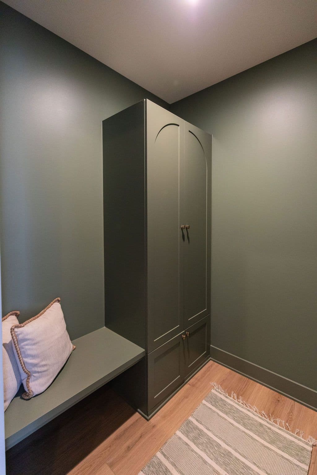 An interior view features a green built-in bench with cushions next to an arched cabinet, complemented by a striped rug and wooden floors, all within a room painted in a similar green hue.