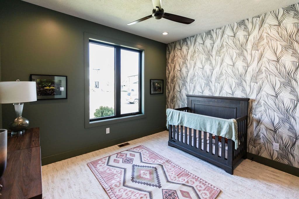 Baby's room with olive green walls, a statement wallpaper accent wall featuring a botanical design, and light wood floors. A dark wood crib sits against the wallpapered wall, complemented by a geometric patterned rug.