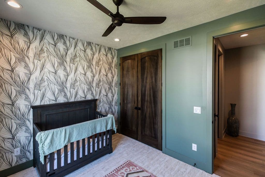 A nursery featuring a dark wood crib with a light green blanket, a wallpapered wall with a black and white leaf pattern, and a ceiling fan. Dark wood doors are visible next to a doorway to an adjacent room with wood flooring.