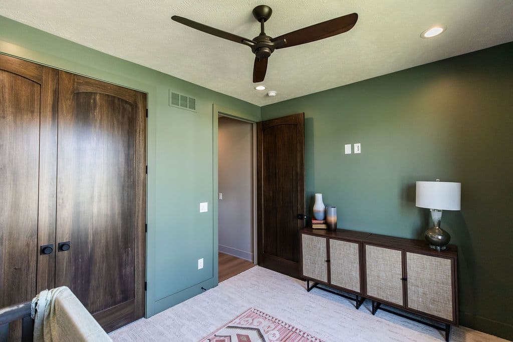 Bedroom with a ceiling fan, a wooden storage cabinet, and a closet with dark brown wooden doors. The walls are painted in a muted green tone, and a doorway leads to another room.
