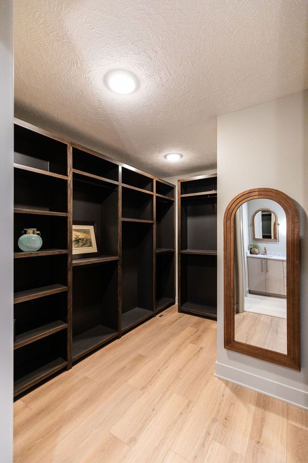 Walk-in closet with built-in dark wood shelving and arch mirror, featuring light wood flooring and view into bathroom. The ceiling has recessed lighting.
