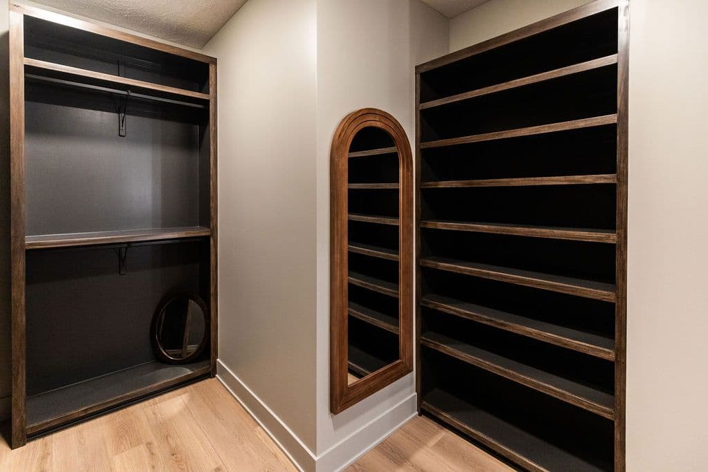 Custom walk-in closet with built-in dark wood shelving and a full-length arched mirror. The closet features light wood flooring and neutral-toned walls.