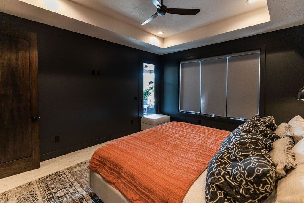 Bedroom features a dark, moody aesthetic with dark brown walls and accents. The room is furnished with a bed covered in a burnt orange comforter, several decorative pillows, and a patterned rug. A door and window are also visible in the room.