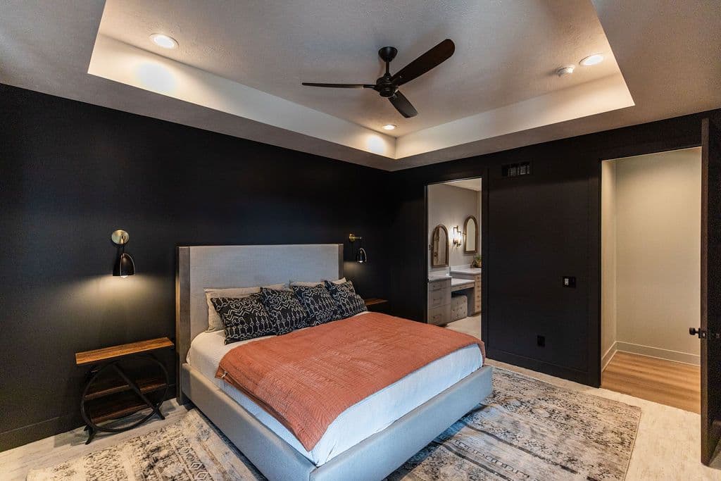 Modern bedroom features a gray upholstered bed with patterned pillows and a salmon-colored throw blanket. The room also includes dark accent walls, a tray ceiling with recessed lighting, and access to an en suite bathroom.