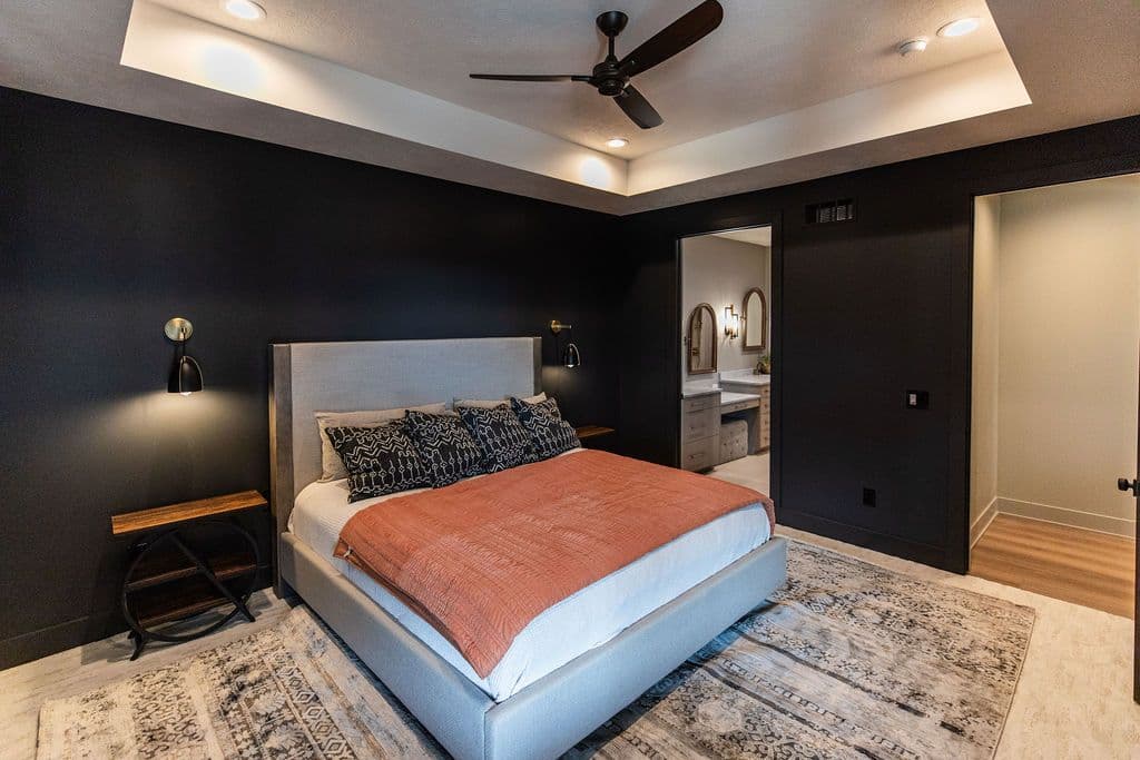 Modern bedroom with dark walls, gray upholstered bed, and textured rug. The room features a tray ceiling with recessed lighting, sconces, and a connecting bathroom.