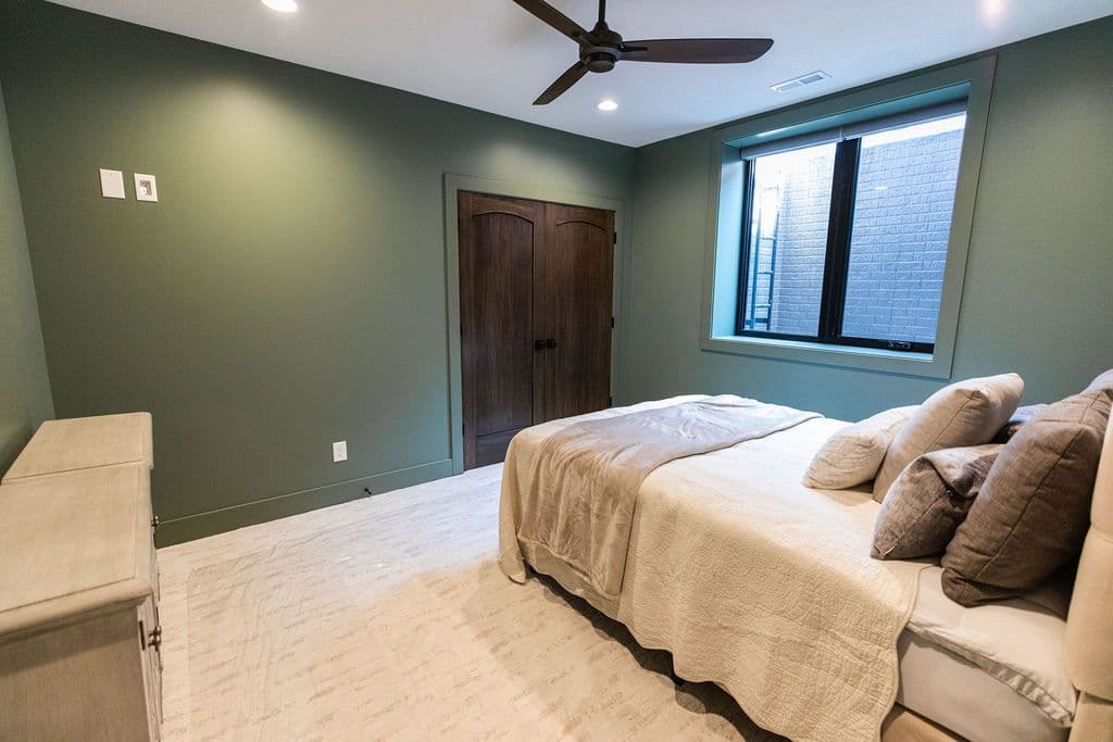 Bedroom with muted green walls features a bed with soft, neutral bedding and decorative pillows, alongside a light-colored dresser. The room also includes a ceiling fan, recessed lighting, and a window overlooking a brick-lined exterior well.