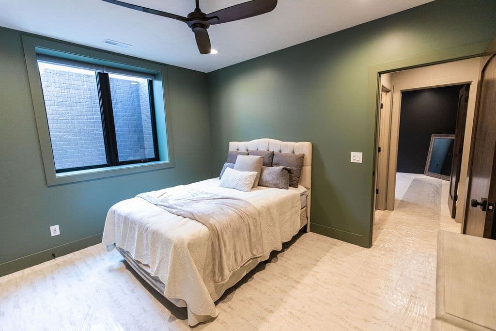 This bedroom features green walls, a beige upholstered bed, and a unique window with a light well. The room has light-colored flooring and leads to an adjacent room with darker walls and a leaning mirror.